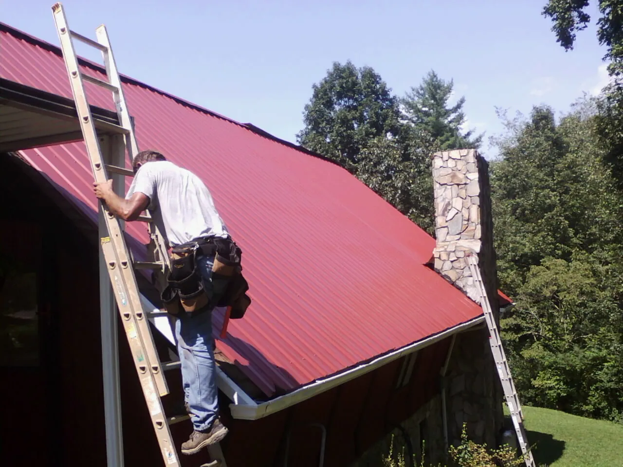 Roofer on a ladder beside a red metal roof and stone chimney — Soffit & Fascia Repair by Roofing Brothers in Sparkill